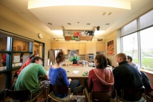 Wide-angle photo of students gathered around a large table at the University of North Dakota's test kitchen. A young female chef/instructor is demonstrating something with a blender toward the back of the table. Ceiling holds a mirror for better views of the demonstration.